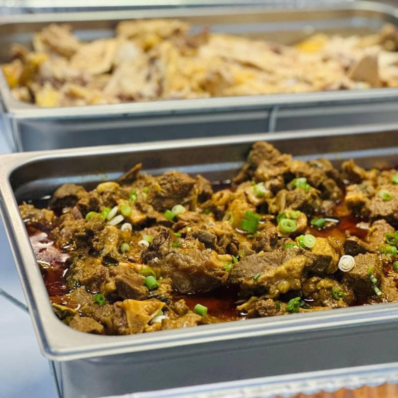 Two buffet trays of Asian-fusion food, with beef and scallions in the foreground.