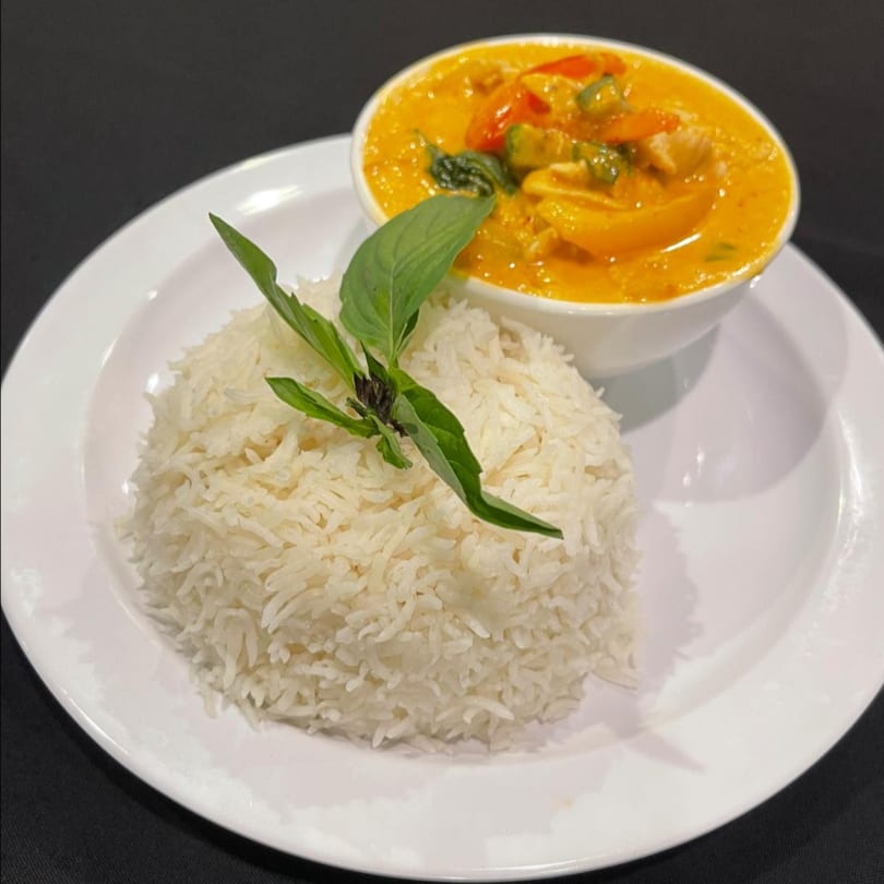 A bowl of yellow curry with chicken and vegetables, served with a side of white rice and basil.