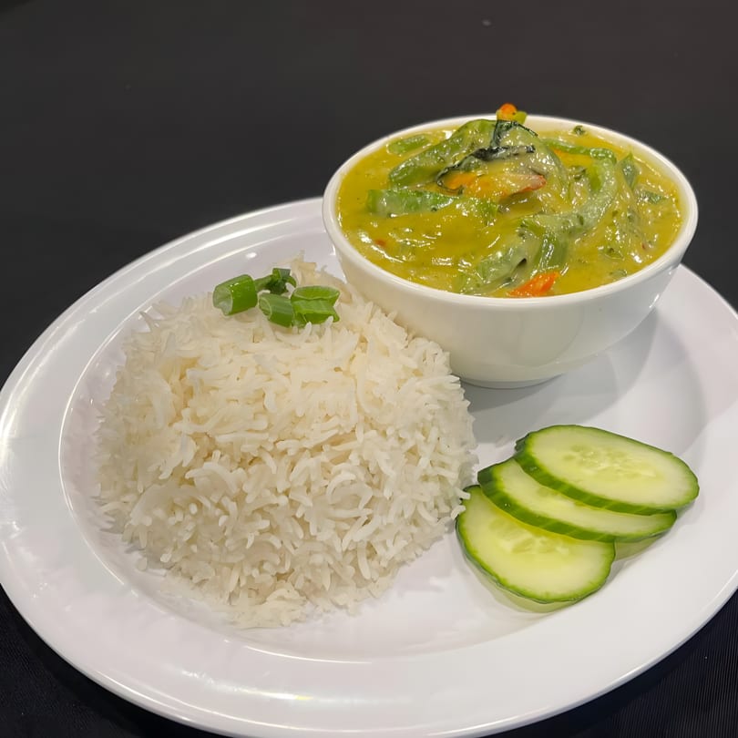 A bowl of green curry with vegetables, white rice, and cucumber slices on a white plate.
