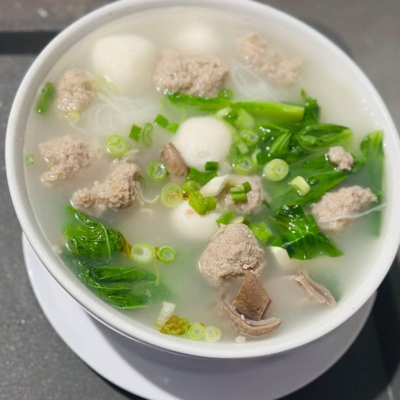 A white bowl of Asian-fusion soup with meatballs, noodles, green onions, and leafy greens.